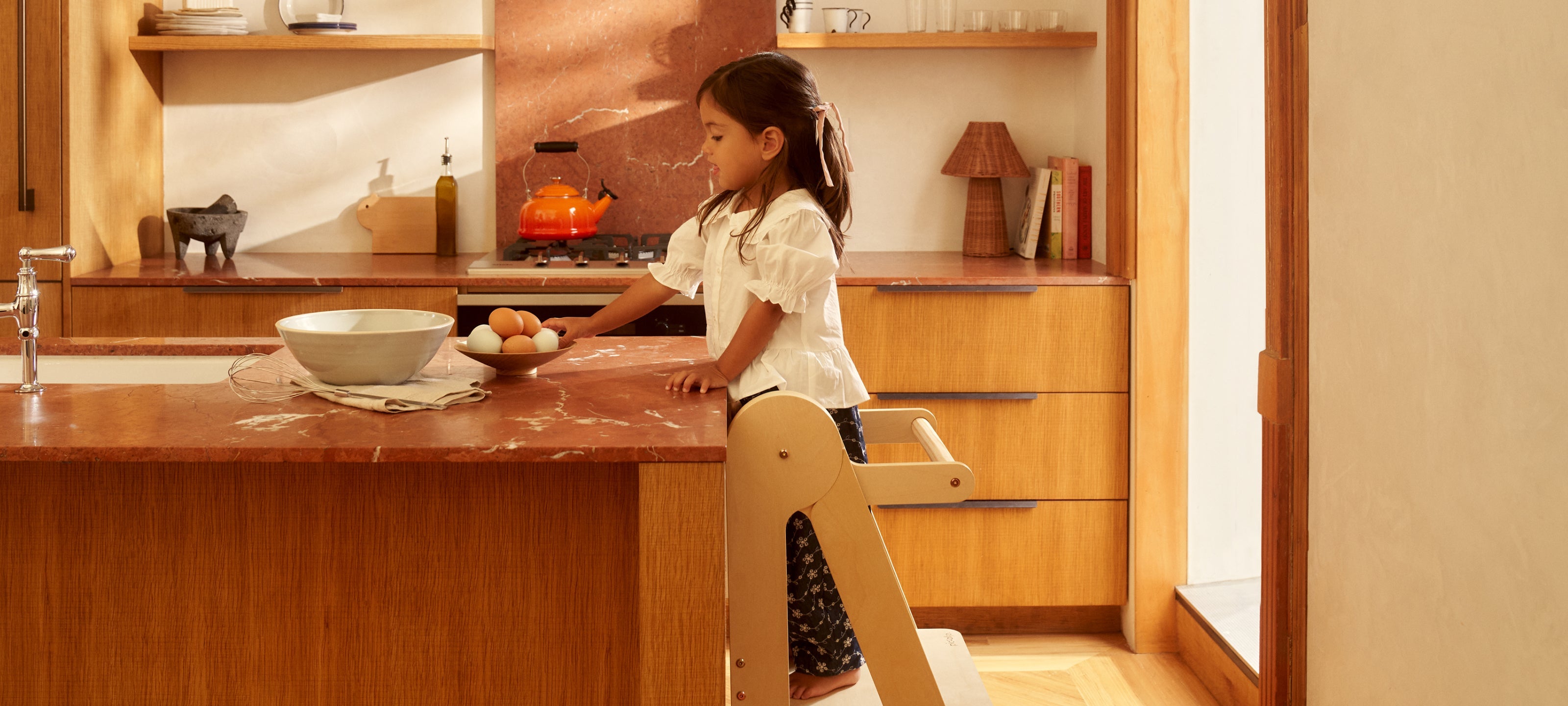 Toddler standing on a foldable toddler tower in birch at a kitchen counter to safely help with cooking and food prep