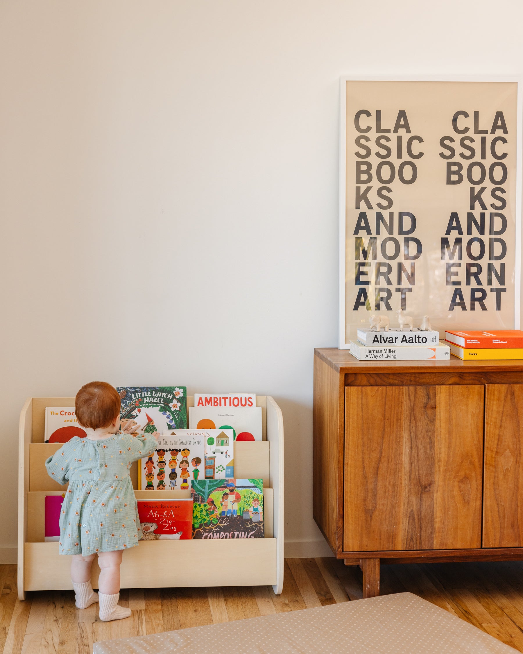 A young girl standing in front of the natural finish Piccalio Montessori bookshelf with colorful books in a living room.