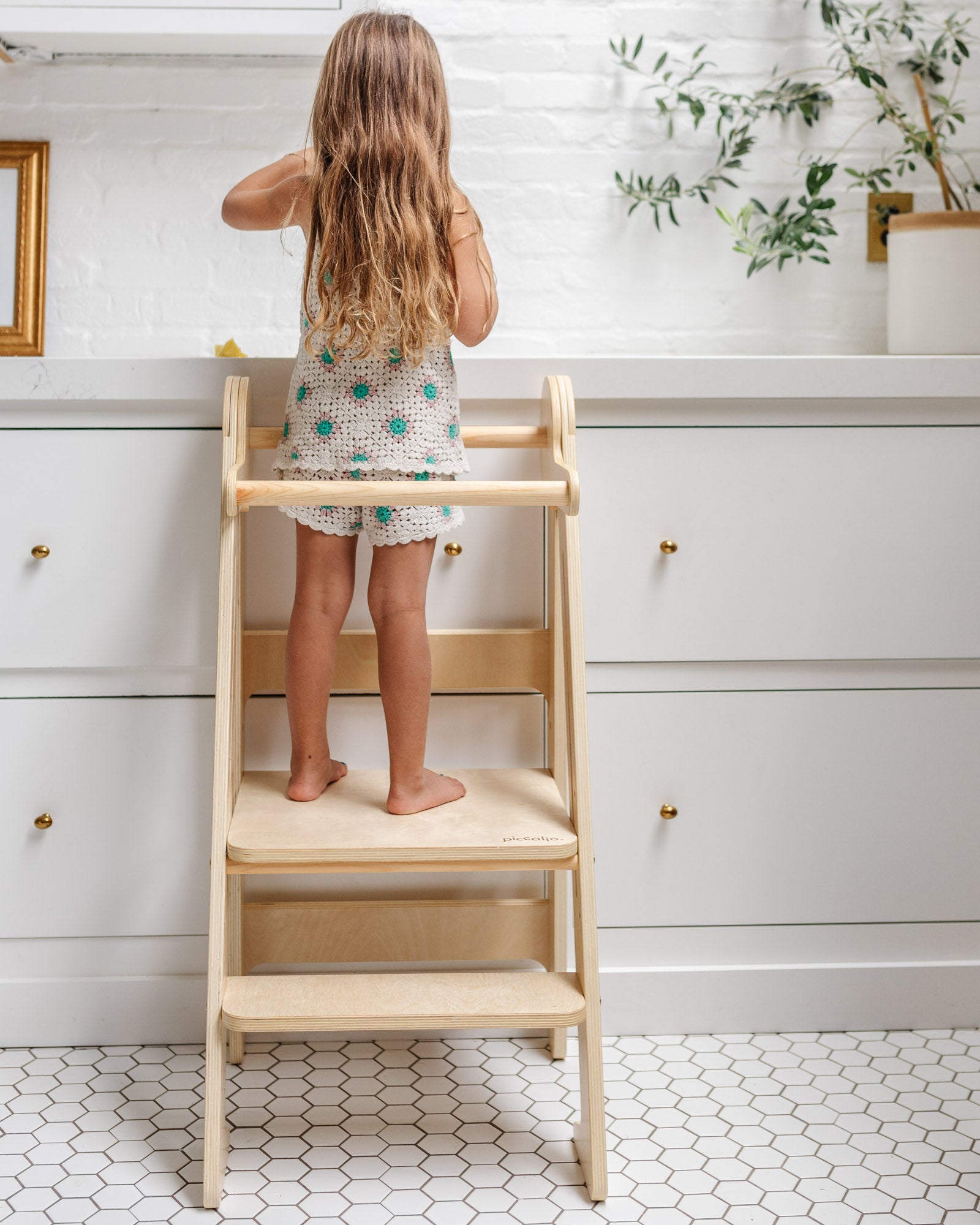 Back view of a child standing in a Piccalio wooden foldable toddler tower to reach the kitchen counter.