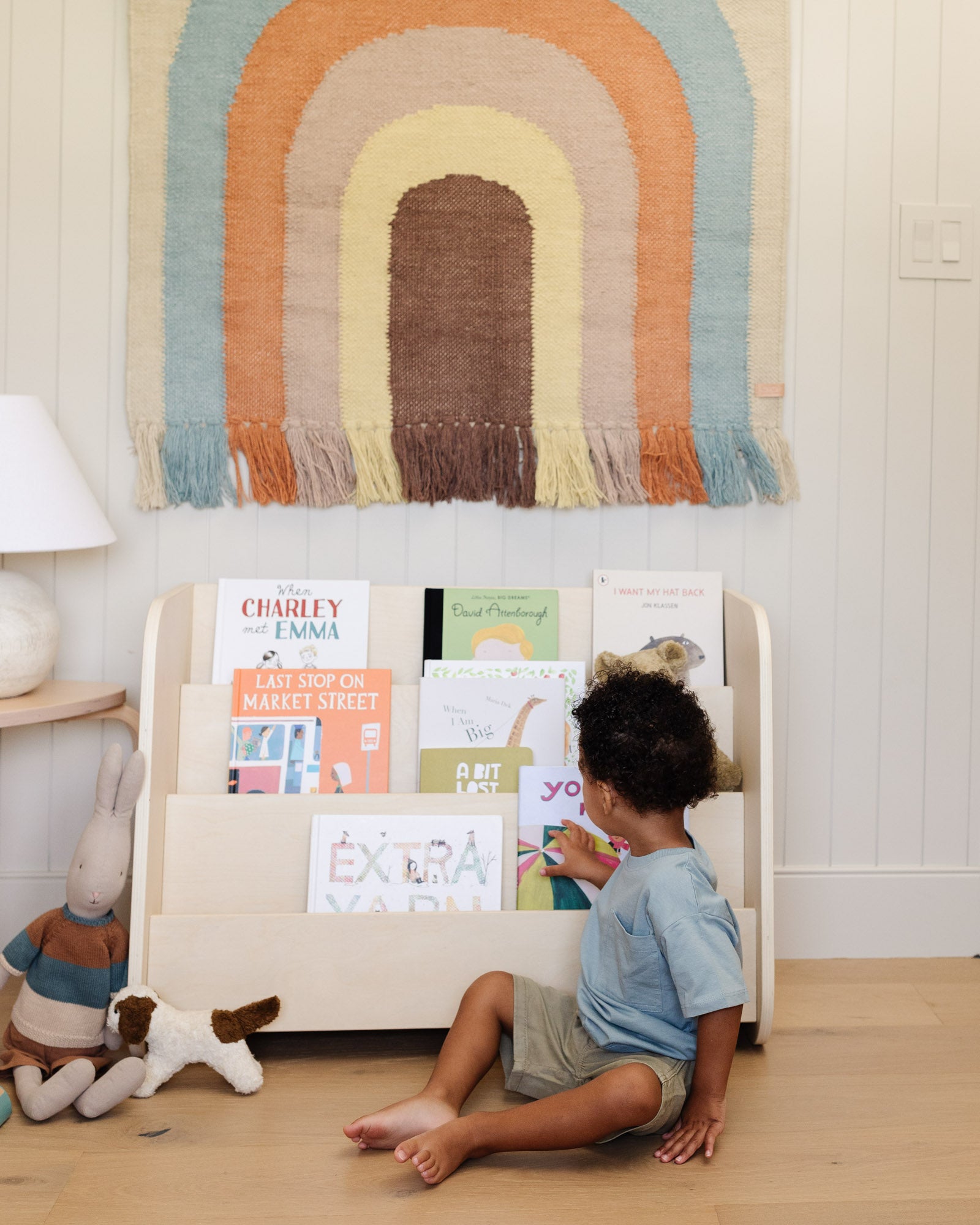 A young boy sitting in front of the natural wood Piccalio Montessori bookshelf, selecting a book from the forward-facing tiers.