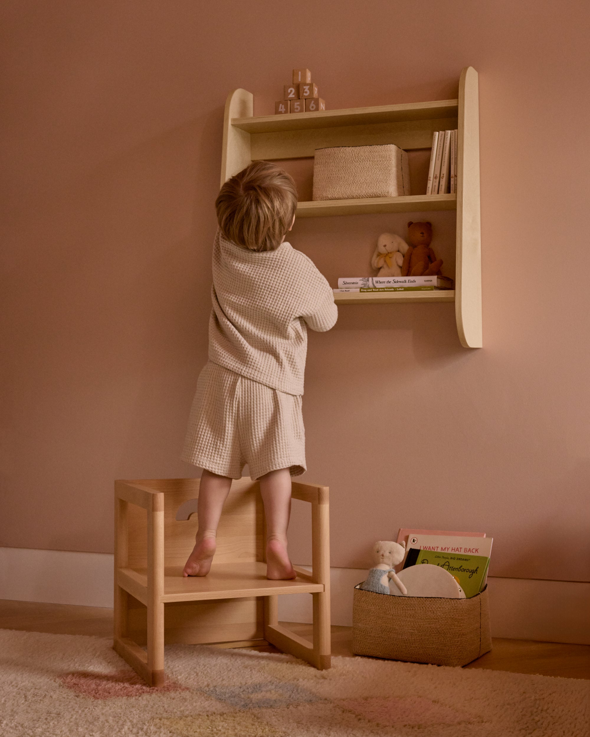 Toddler reaching for books on a Piccalio birch wall shelf while standing on a wooden Montessori Piccalio chair.