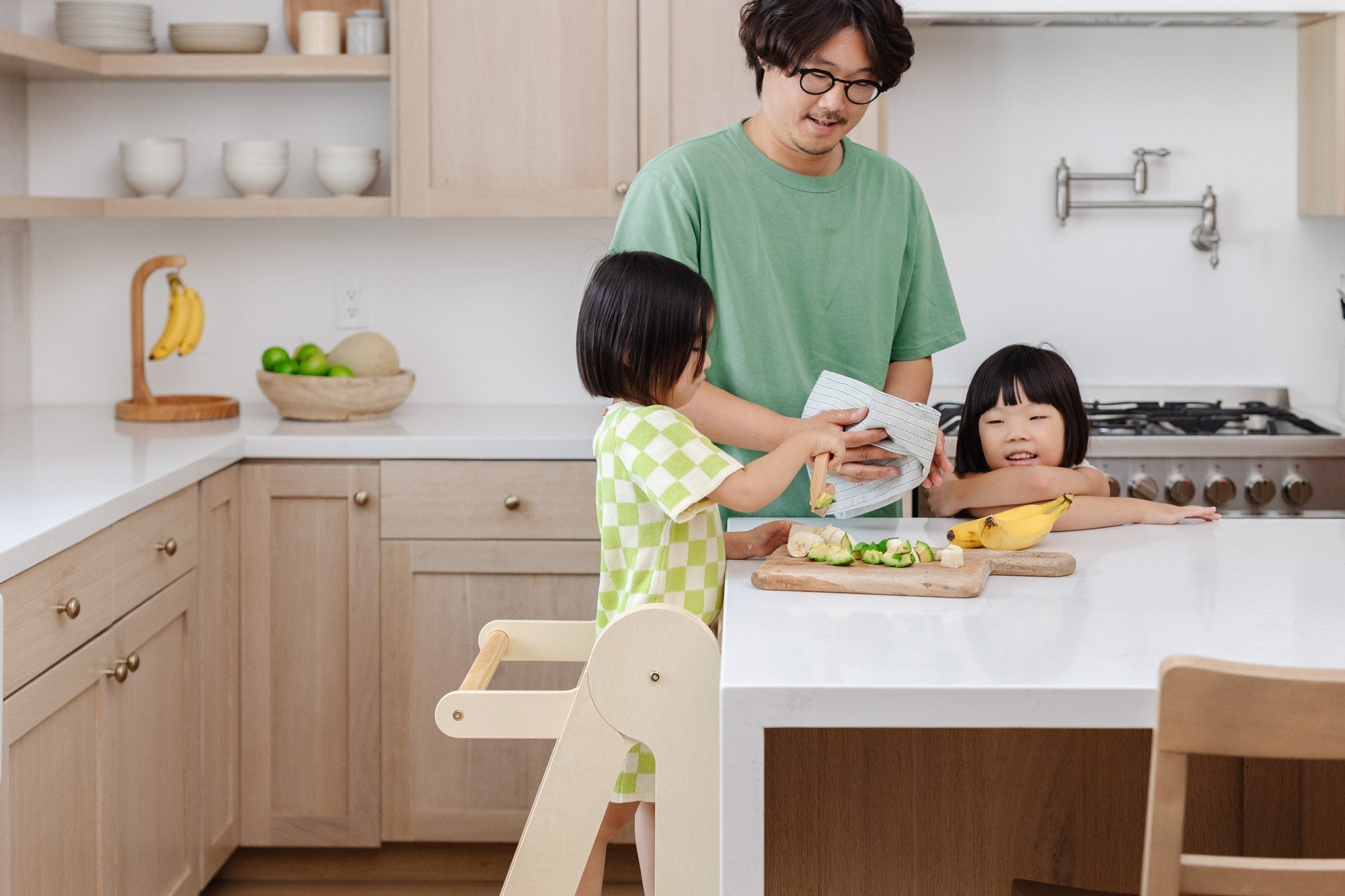 Child using a foldable toddler tower to prepare food with father and sibling in a modern kitchen
