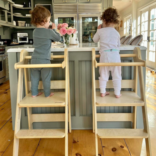 Twins using two foldable toddler towers made of natural birch wood in the kitchen