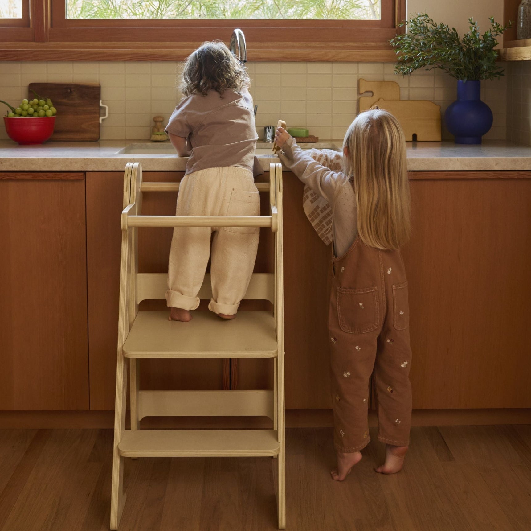 Two children at sink, one using a foldable learning tower to wash hands