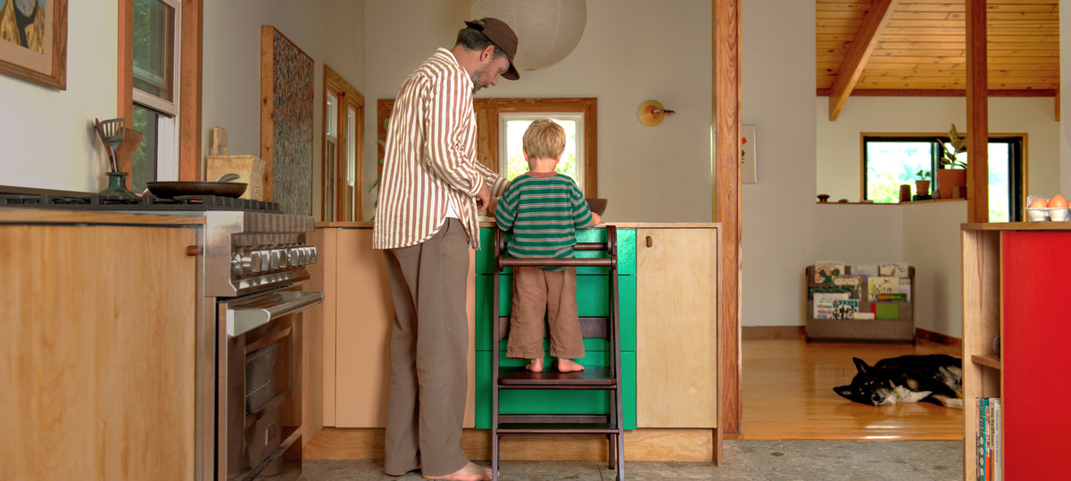 Toddler and father using the Piccalio Foldable Toddler Tower in a walnut stain to prepare food in the kitchen.