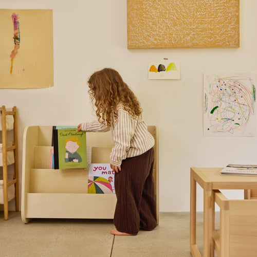 Child returning book to the Montessori bookshelf in natural wood with a table and chair in the foreground.