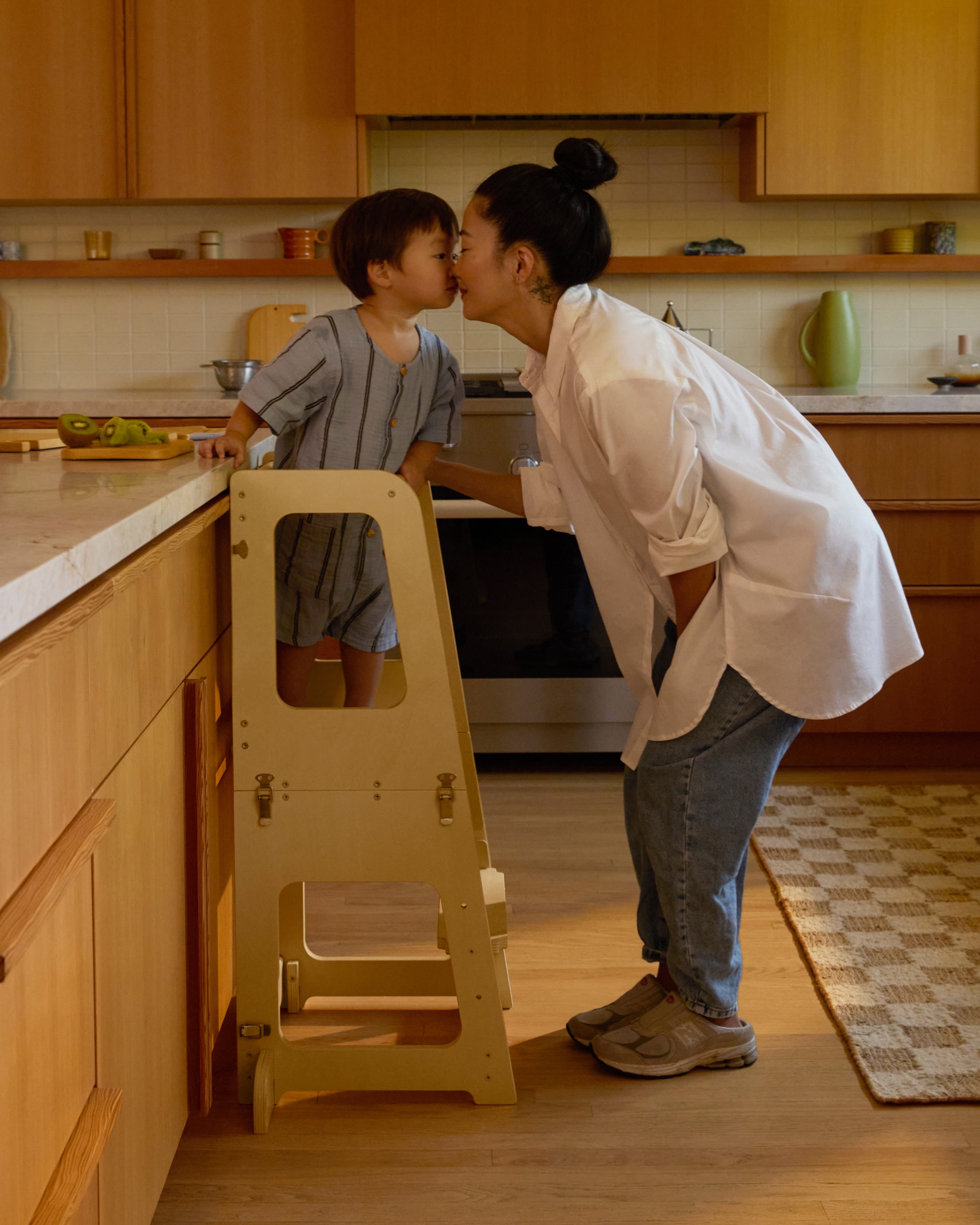 Piccalio Convertible Toddler Tower in natural wood with a toddler standing on the tower with the mother by his side.