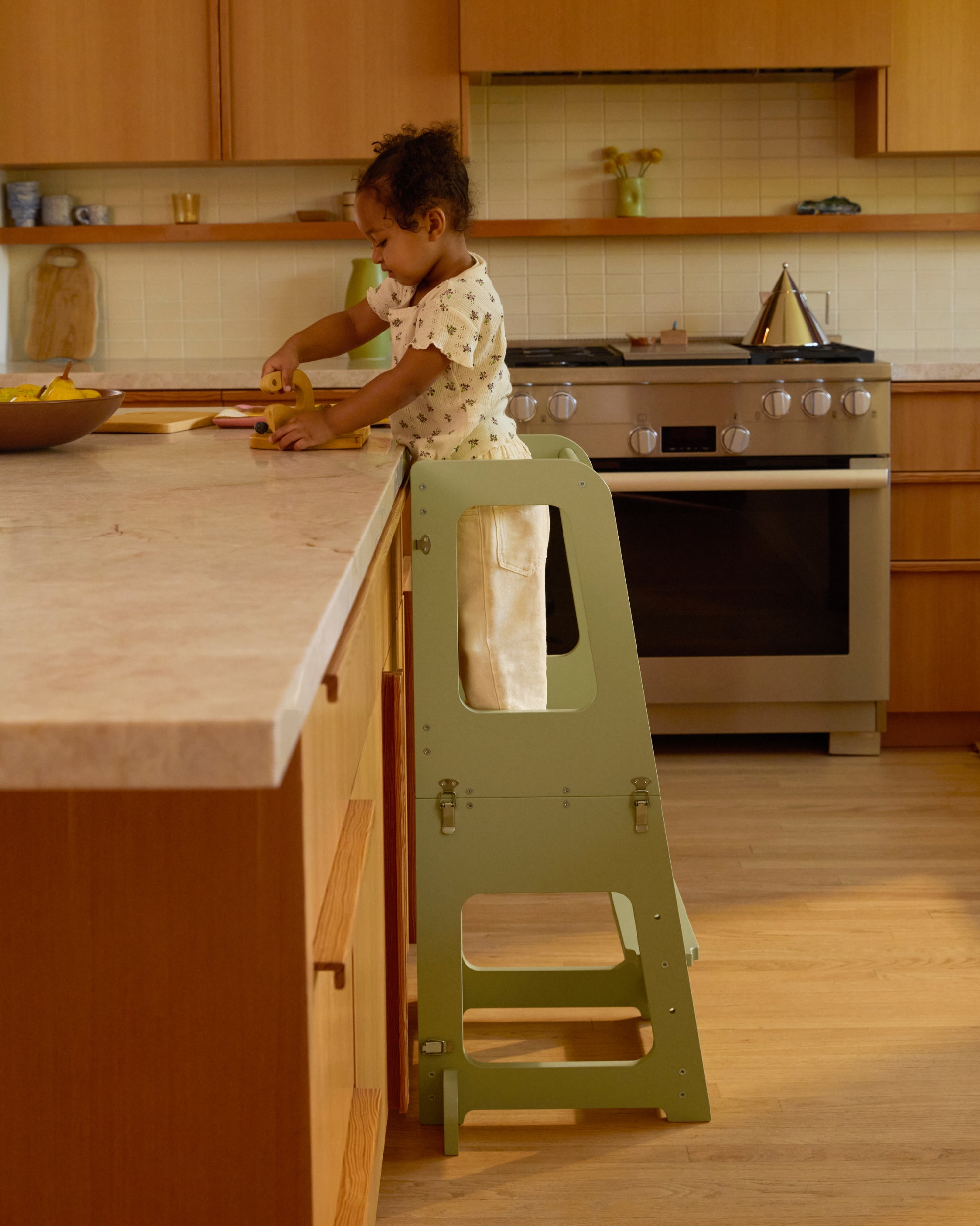 Piccalio convertible toddler tower in sage green with a toddler standing on the tower cutting fruit in the kitchen.