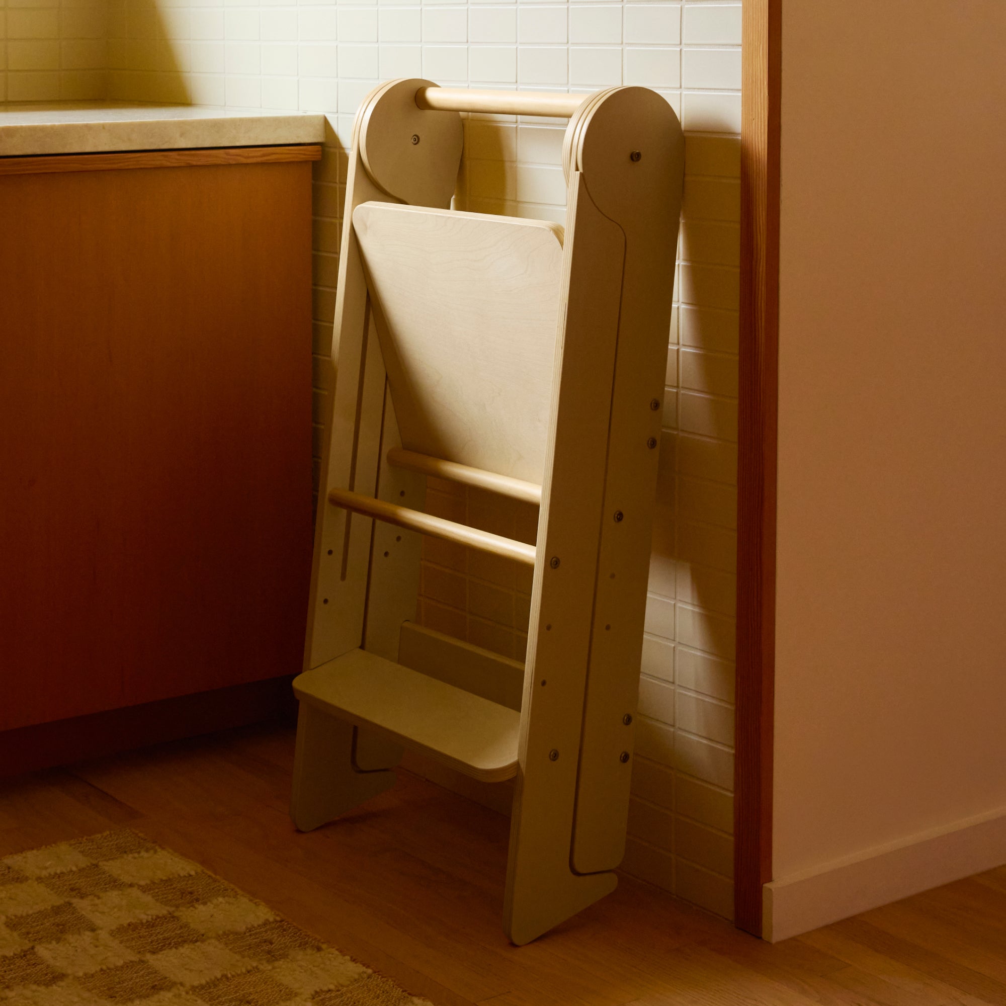 Piccalio Foldable Toddler Tower in natural wood leaning against wall in a kitchen.