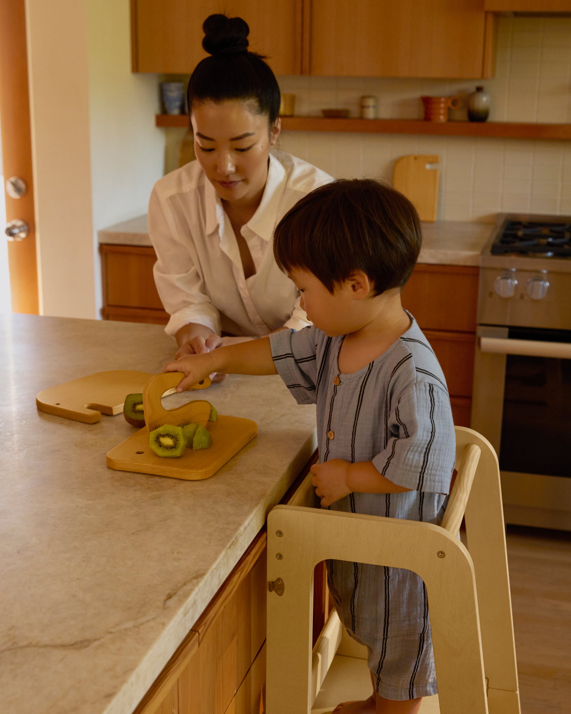 Piccalio convertible toddler tower with a toddler using a wooden knife a cutting fruit with the mother by his side.