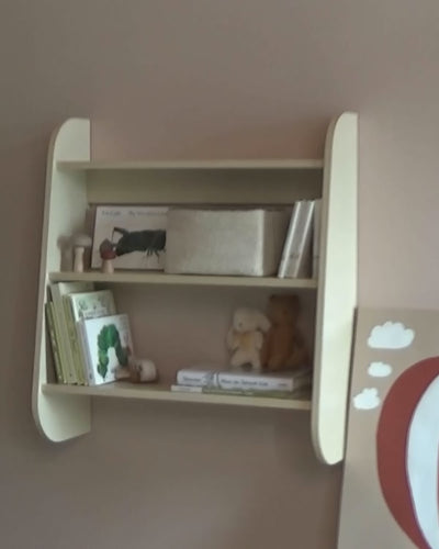 Child arranges wooden number blocks on a Piccalio three-tier birchwood Wall Shelf styled with picture books, a woven basket, and stuffed animals in a Montessori nursery.