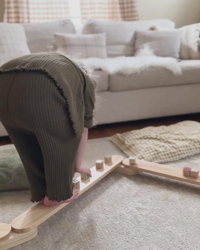 Toddler walks the Piccalio Acrobat Balance Beam in a zigzag setup with wooden blocks while mom assembles the modular Montessori balance beam for kids in a living room.