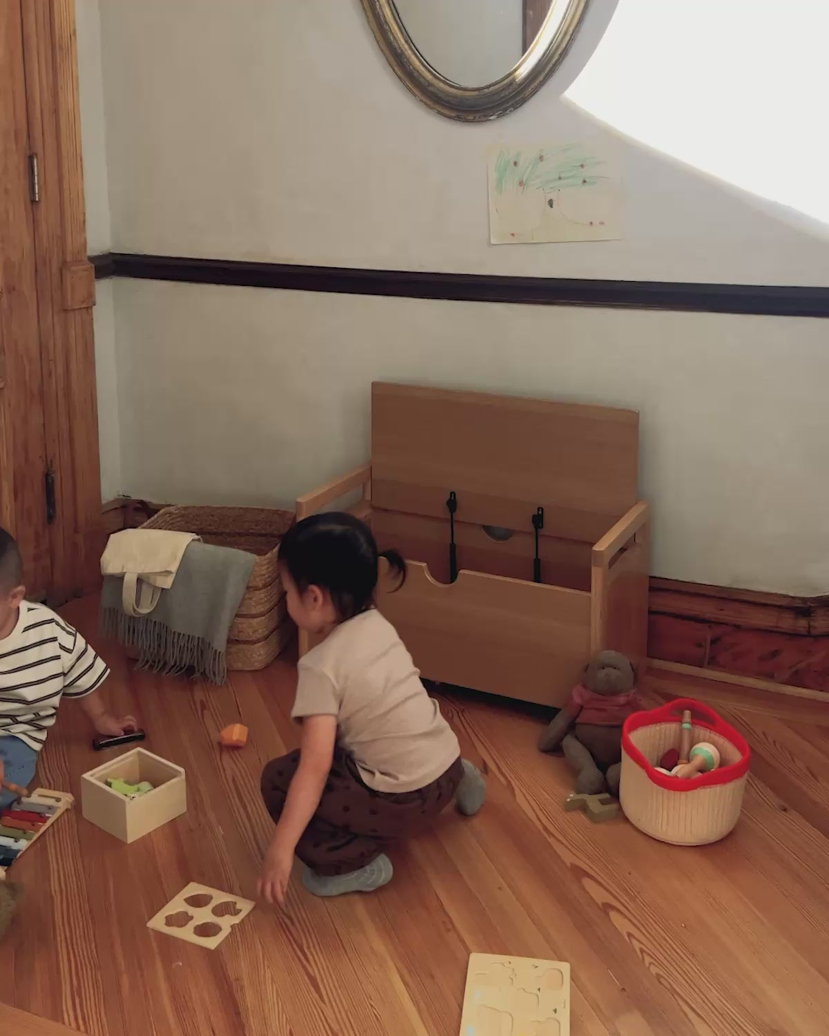 Toddlers put toys away inside the Piccalio Toy Box, a solid oak wooden toy chest for kids with a soft-close safety lid that supports independent cleanup in a Montessori playroom.